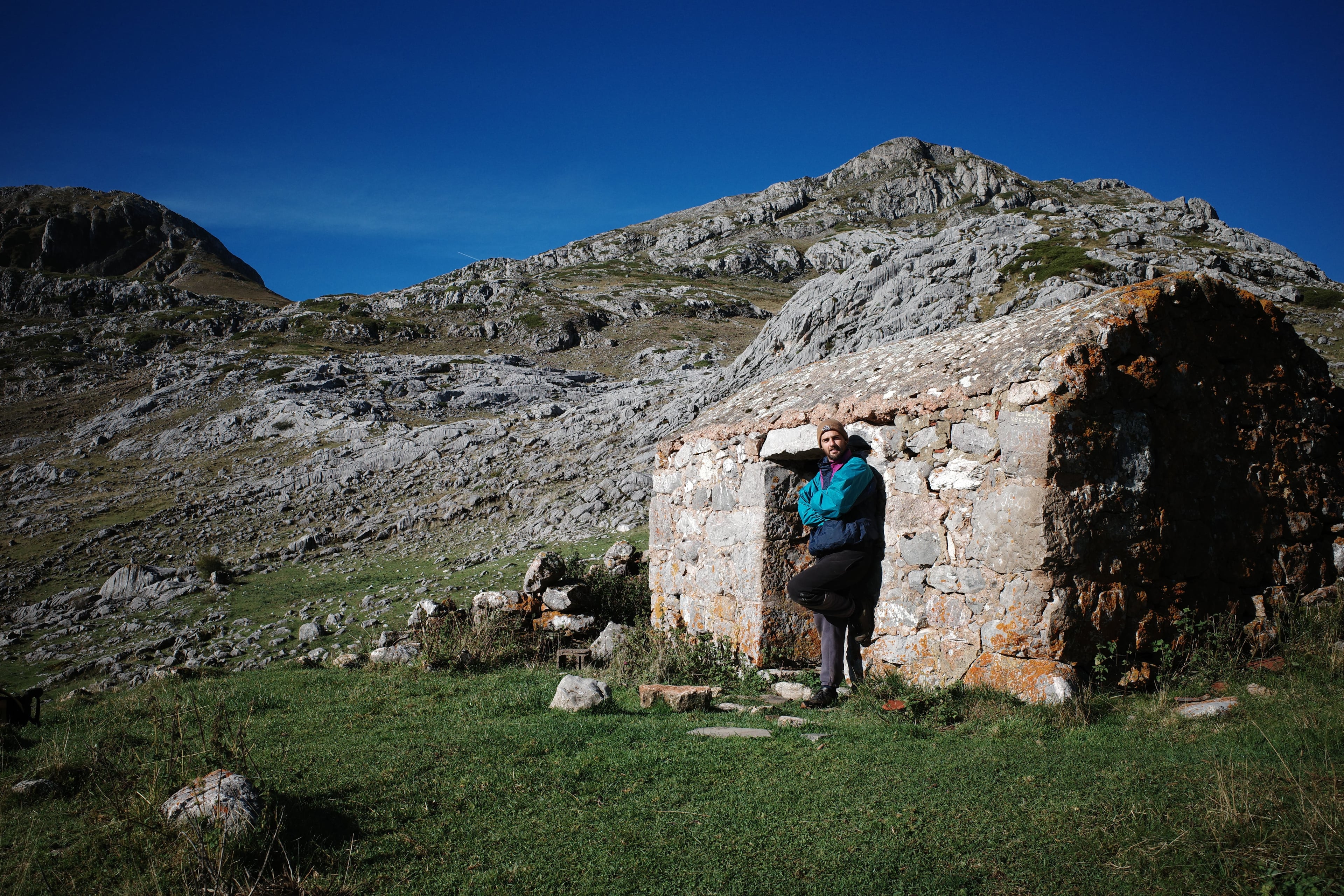 Pato Turri leaning against a stone building on a mountainside, under a clear sky
