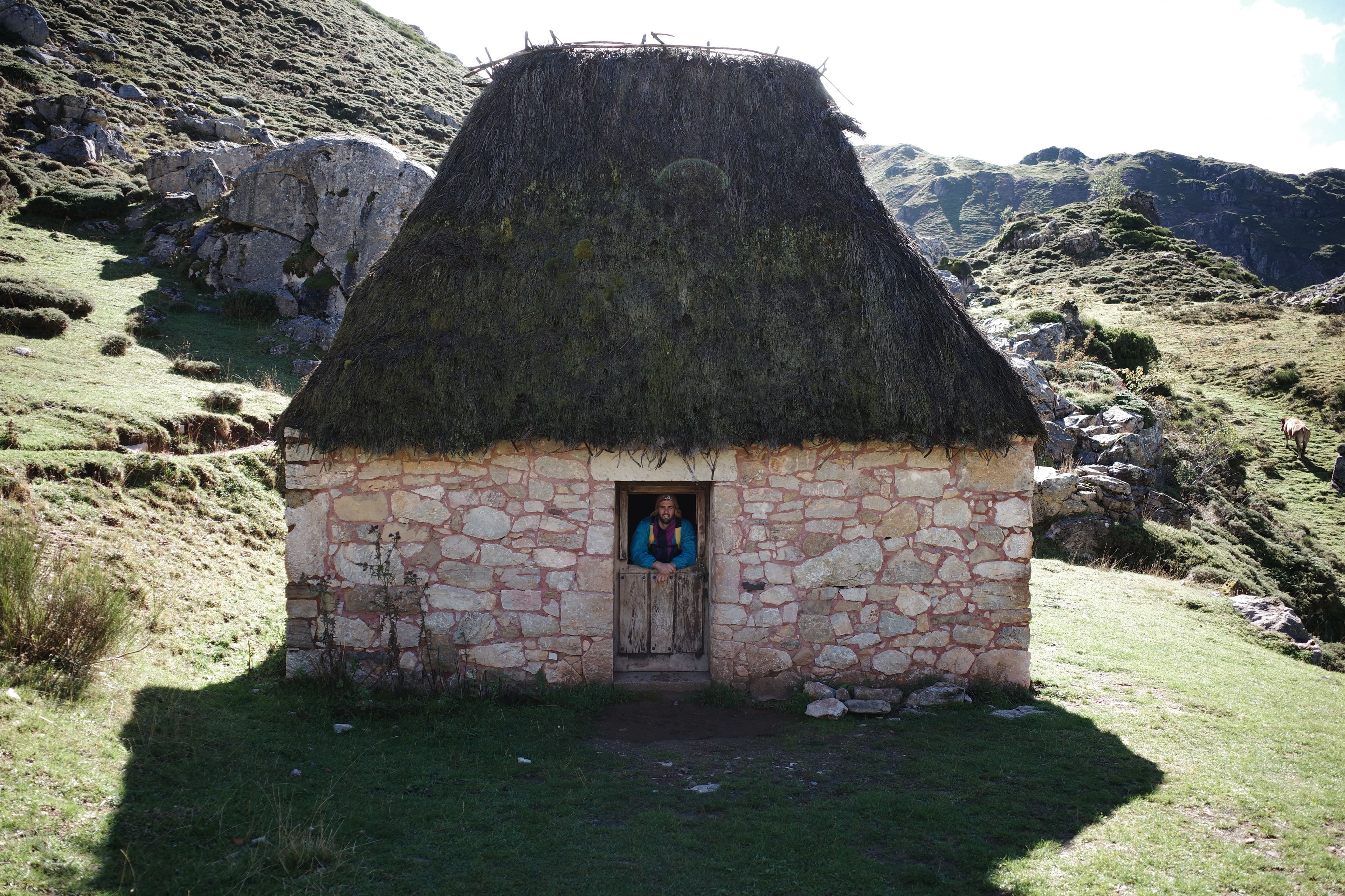 Pato Turri leaning out of the doorway of a stone hut with a thatched roof, in a mountain valley