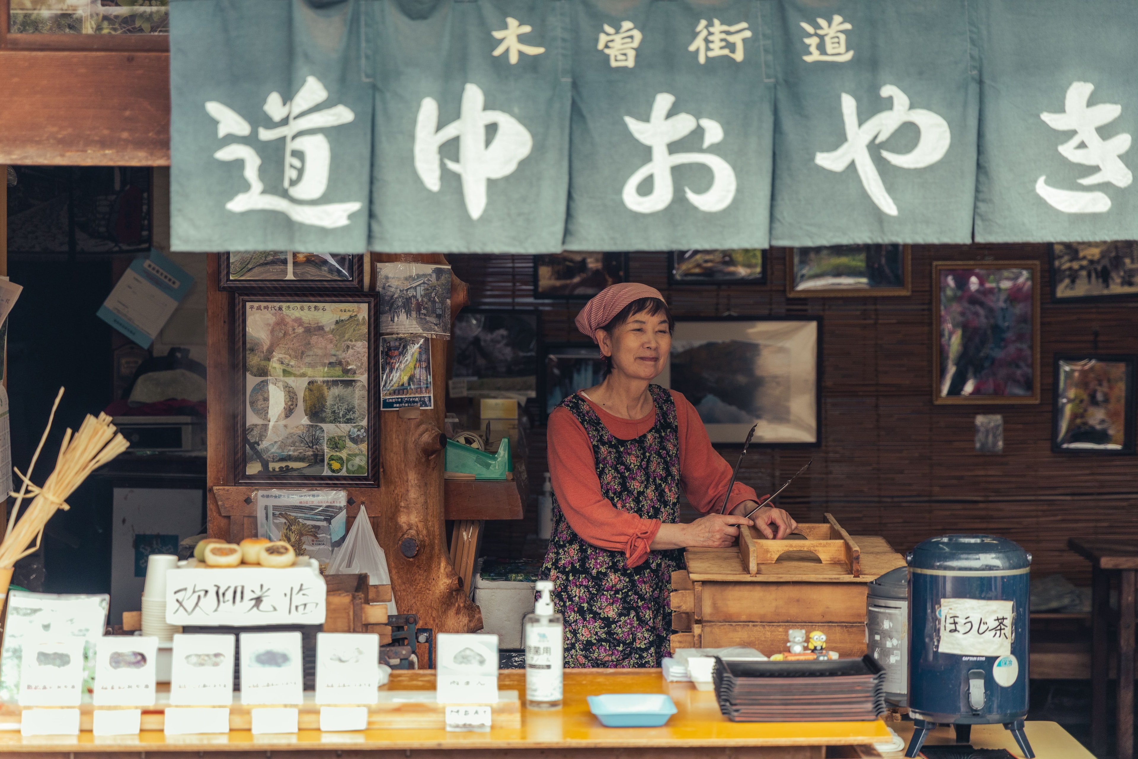 Una mujer atiende un puesto de comida de madera al borde del camino en Japon, bajo una cortina de tela colgada, entre fotografias enmarcadas y un termo de te